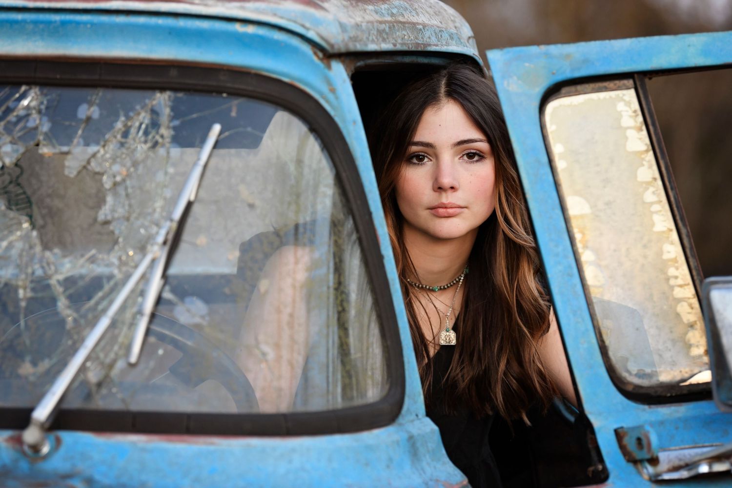 A woman with dark hair peers out from the driver's side window of a vintage blue car.