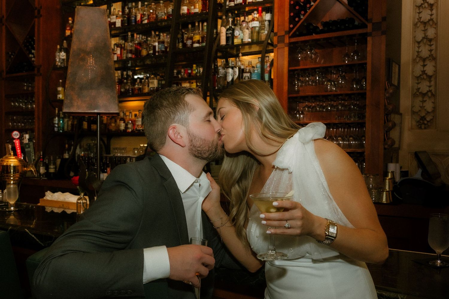 A couple shares a romantic kiss at an elegant bar with bottles of liquor displayed on shelves in the background.