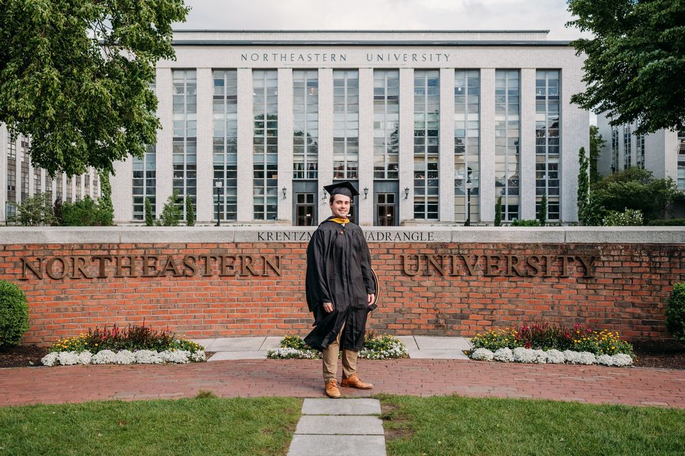 Graduate stands in front of Northeastern University's brick entrance sign wearing academic regalia on graduation day.