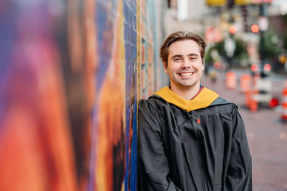 Graduate in black gown and yellow hoodie smiling while leaning against colorful wall on city street.