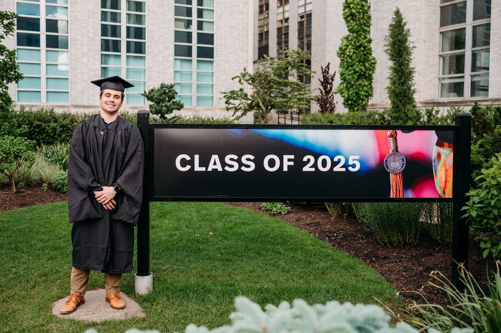 Graduate in cap and gown standing beside Class of 2025 sign on university campus.