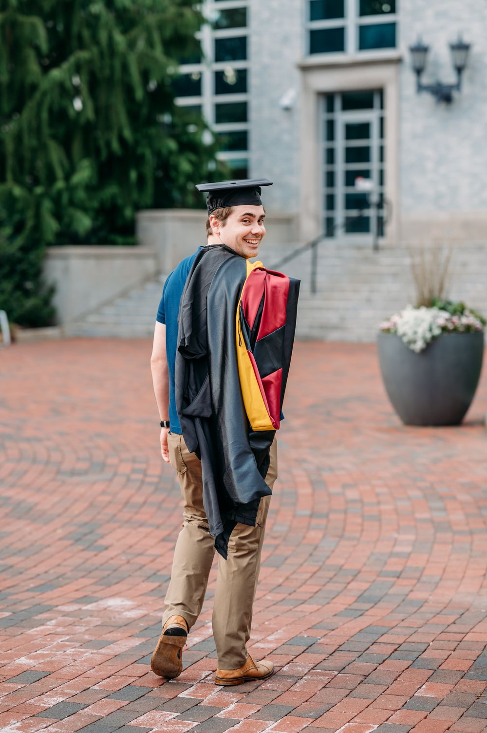 Graduate in cap and gown stands on brick courtyard outside academic building with decorative sash and brown shoes.
