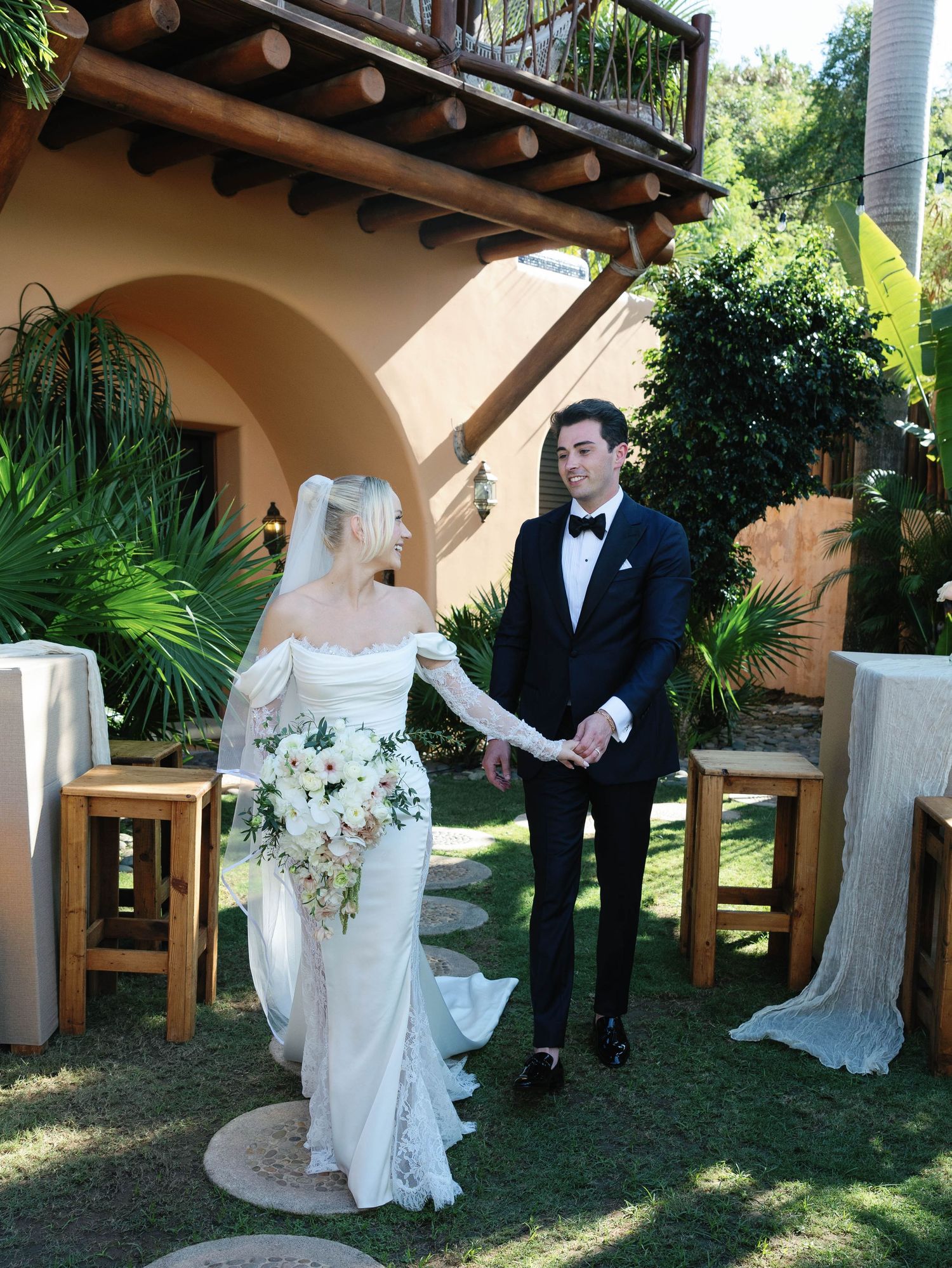 Wedding couple stands under Spanish-style archway surrounded by lush tropical landscaping.