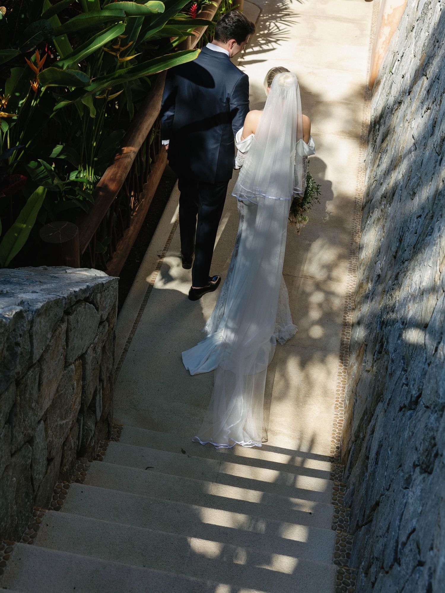 A wedding couple descends stone stairs with flowing veil trailing behind on a sunny day.