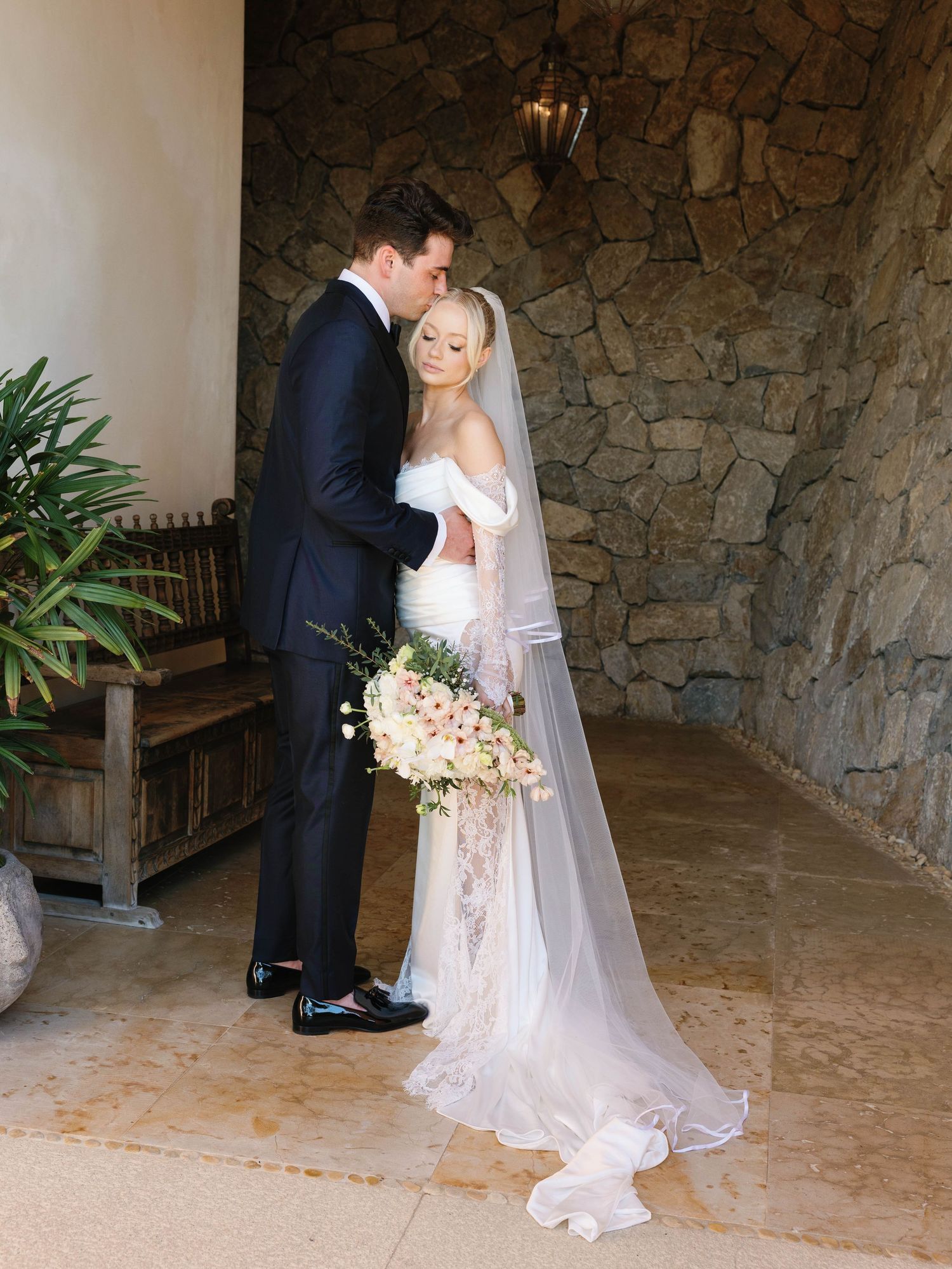 A romantic wedding moment against a rustic stone wall with blush florals and long veil.