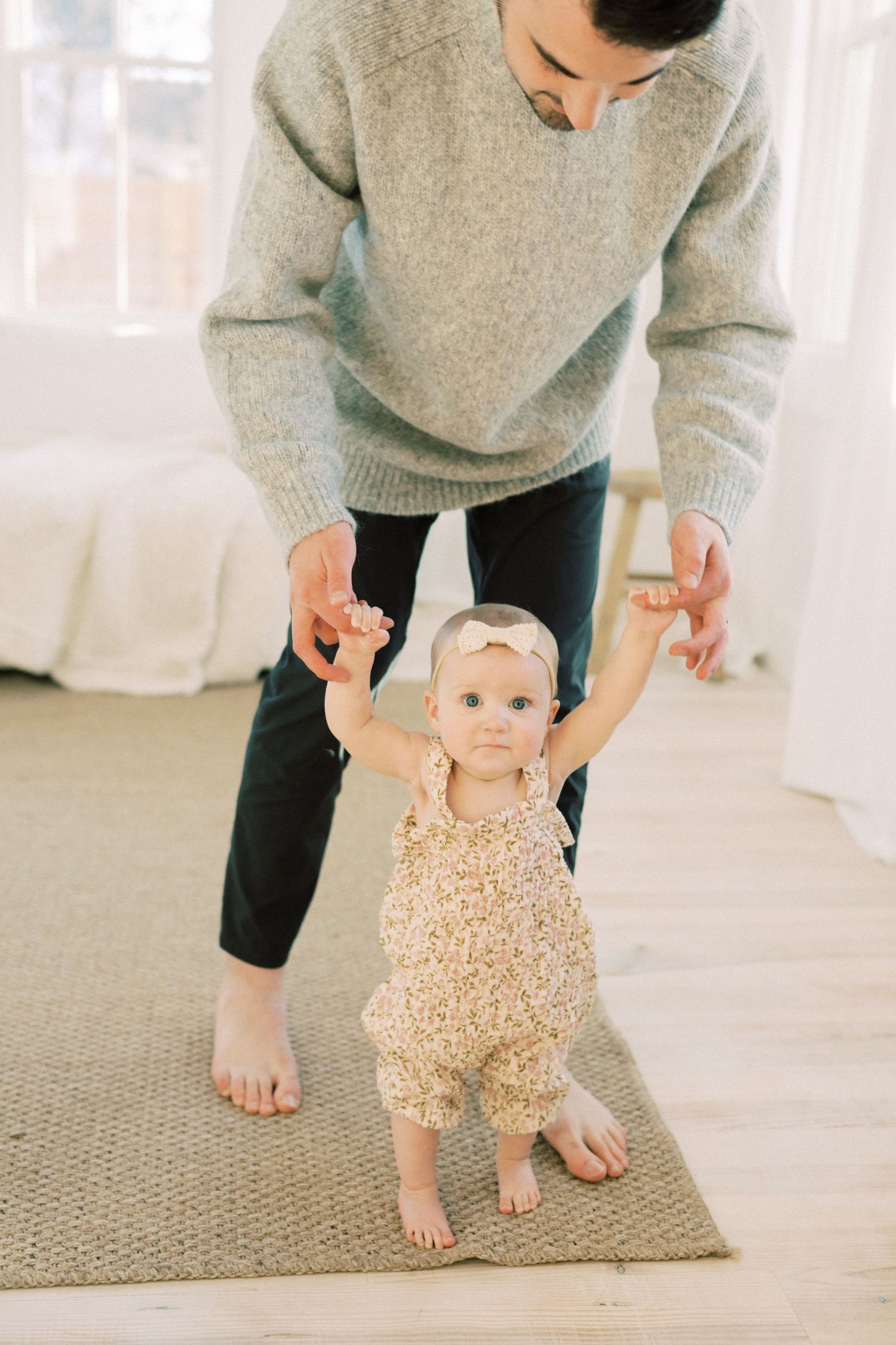 A baby in a floral romper takes first steps while being supported on a gray rug in a bright room.