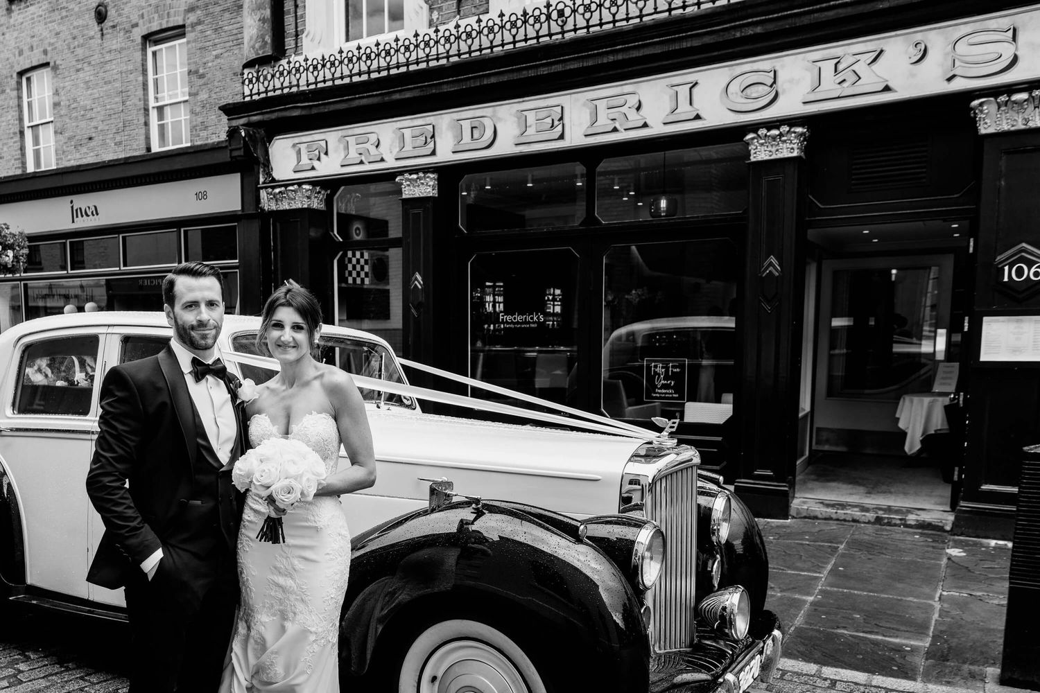 Classic vintage white Rolls Royce wedding car parked in front of Frederick's storefront in black and white photograph.