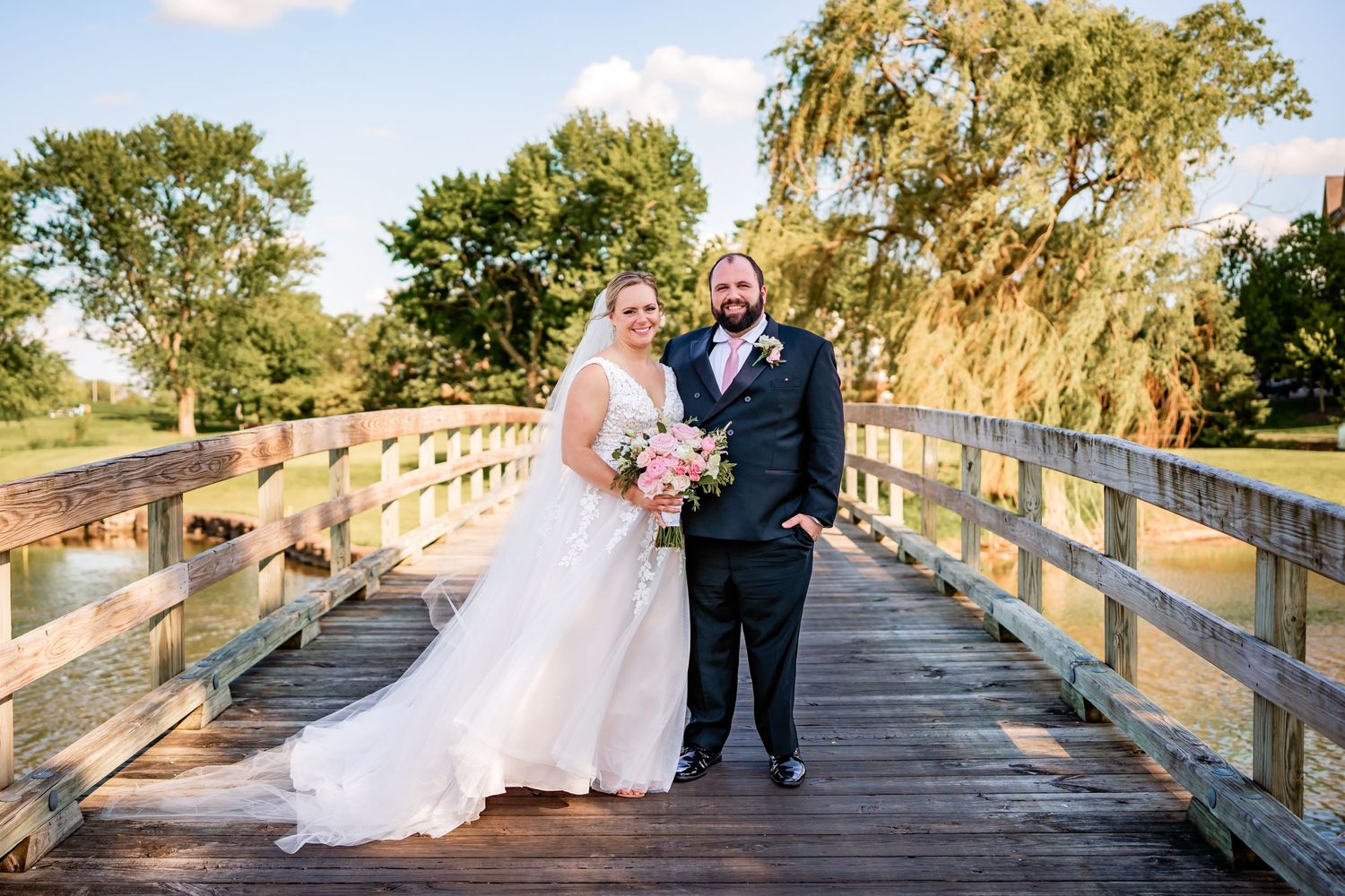 A wedding couple poses on a rustic wooden bridge surrounded by lush greenery.