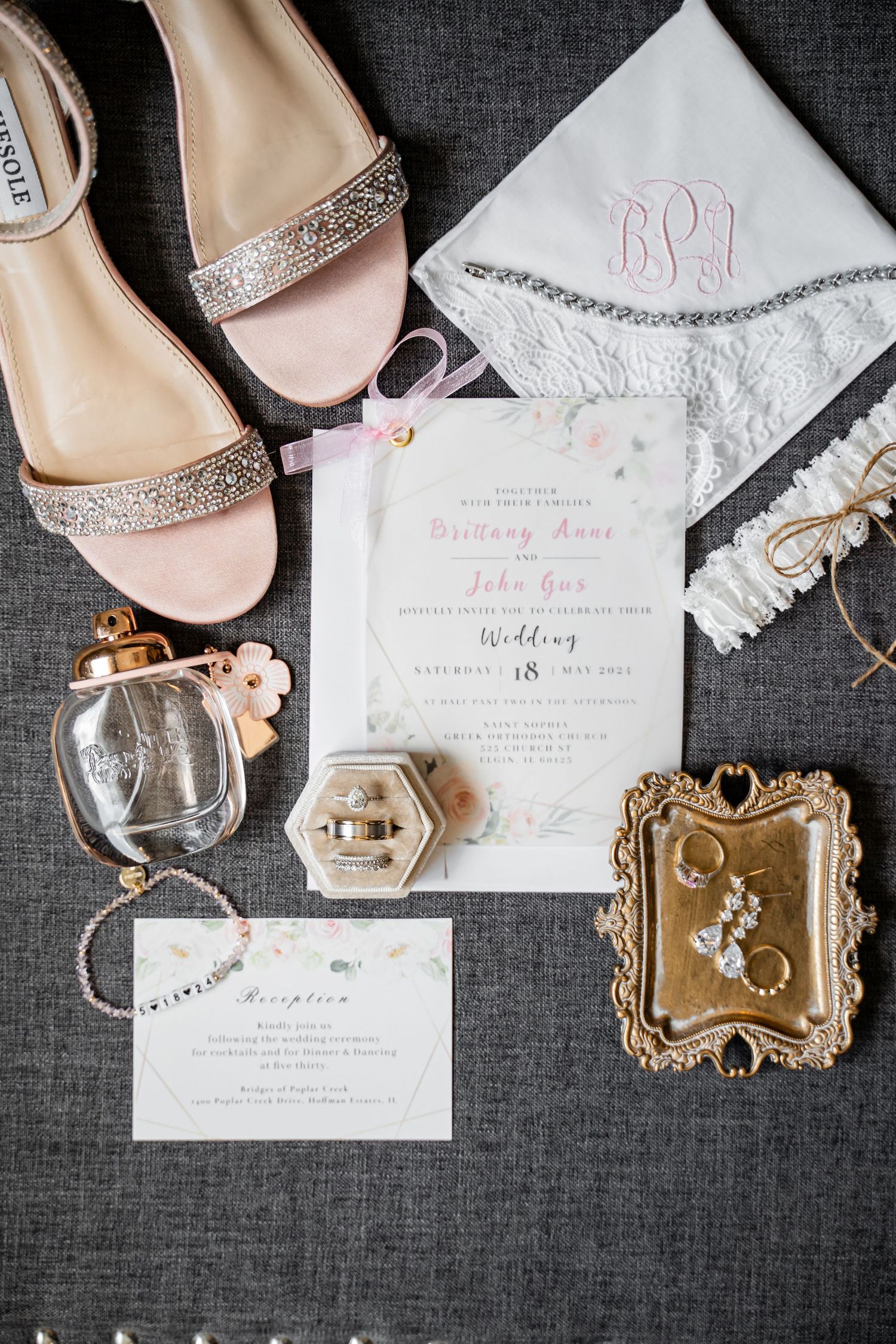 Wedding details flatlay with sparkly shoes, invitation, monogrammed handkerchief and jewelry on grey fabric.