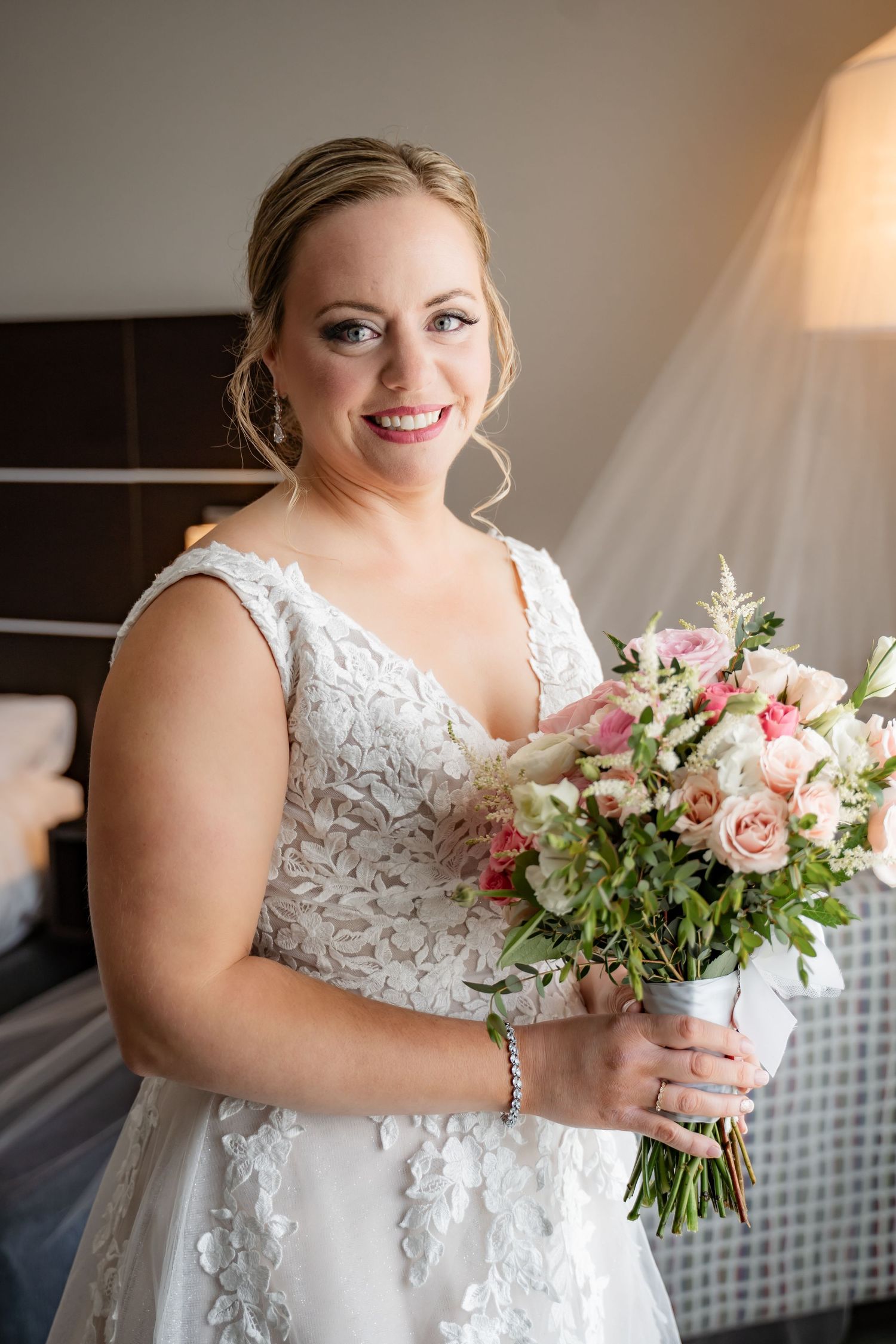 Bride in lace wedding dress holds pink and white bouquet while smiling.