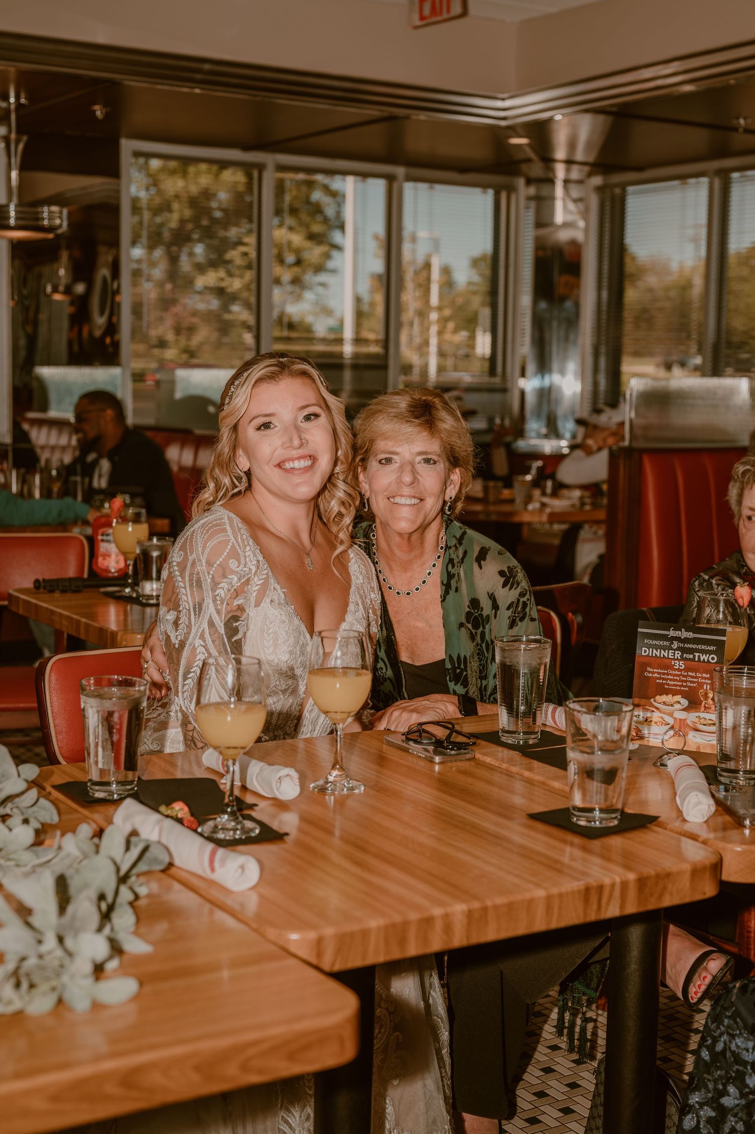 Bride and mom enjoying drinks and conversation at a diner restaurant with wooden tables in warm ambient lighting by Samantha Campbell