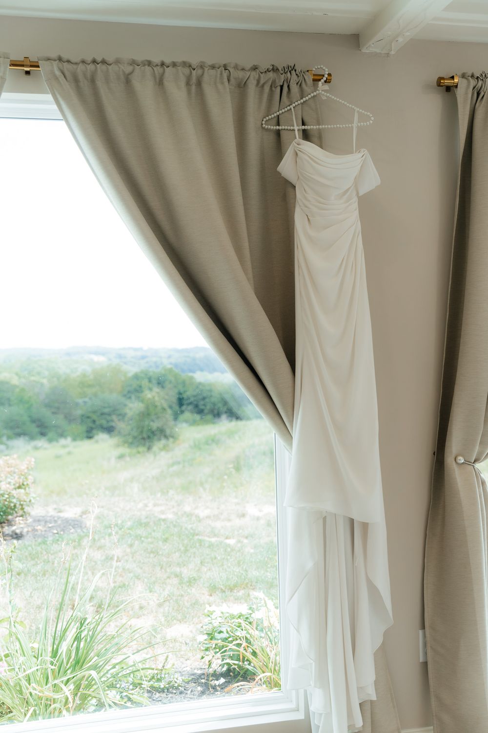 White wedding dress hanging by a window with taupe curtains and scenic green view outside.