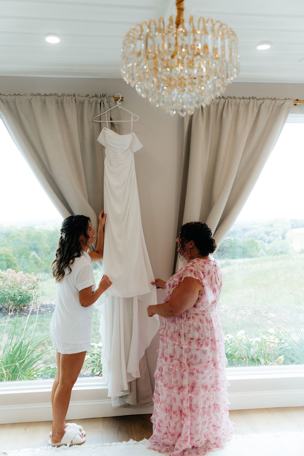 Two people admiring an elegant white wedding gown hung by a window with a crystal chandelier.