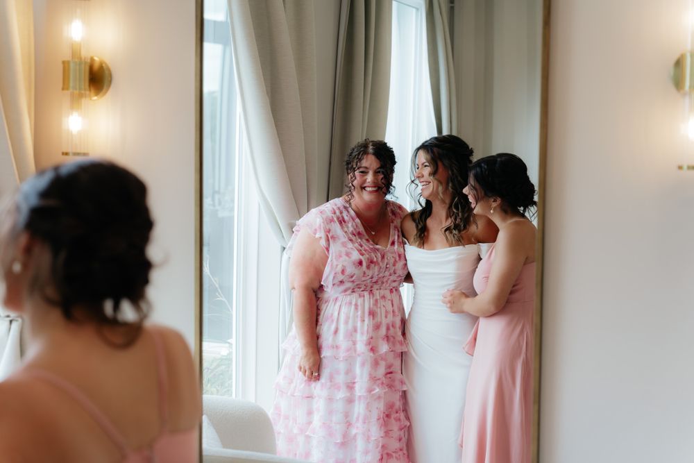 Three women share a special moment in an elegant room with soft lighting and white curtains as they prepare for a wedding.