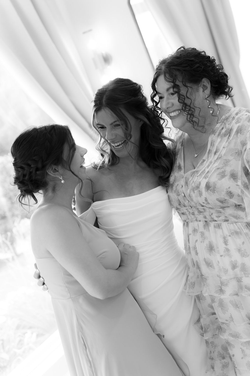 Three women in wedding attire share a candid laughing moment together in black and white photography.