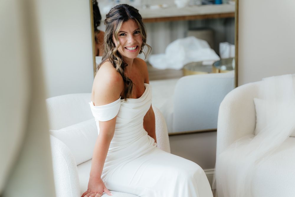 Person wearing an elegant white off-shoulder wedding dress sits on white furniture in a bright room.