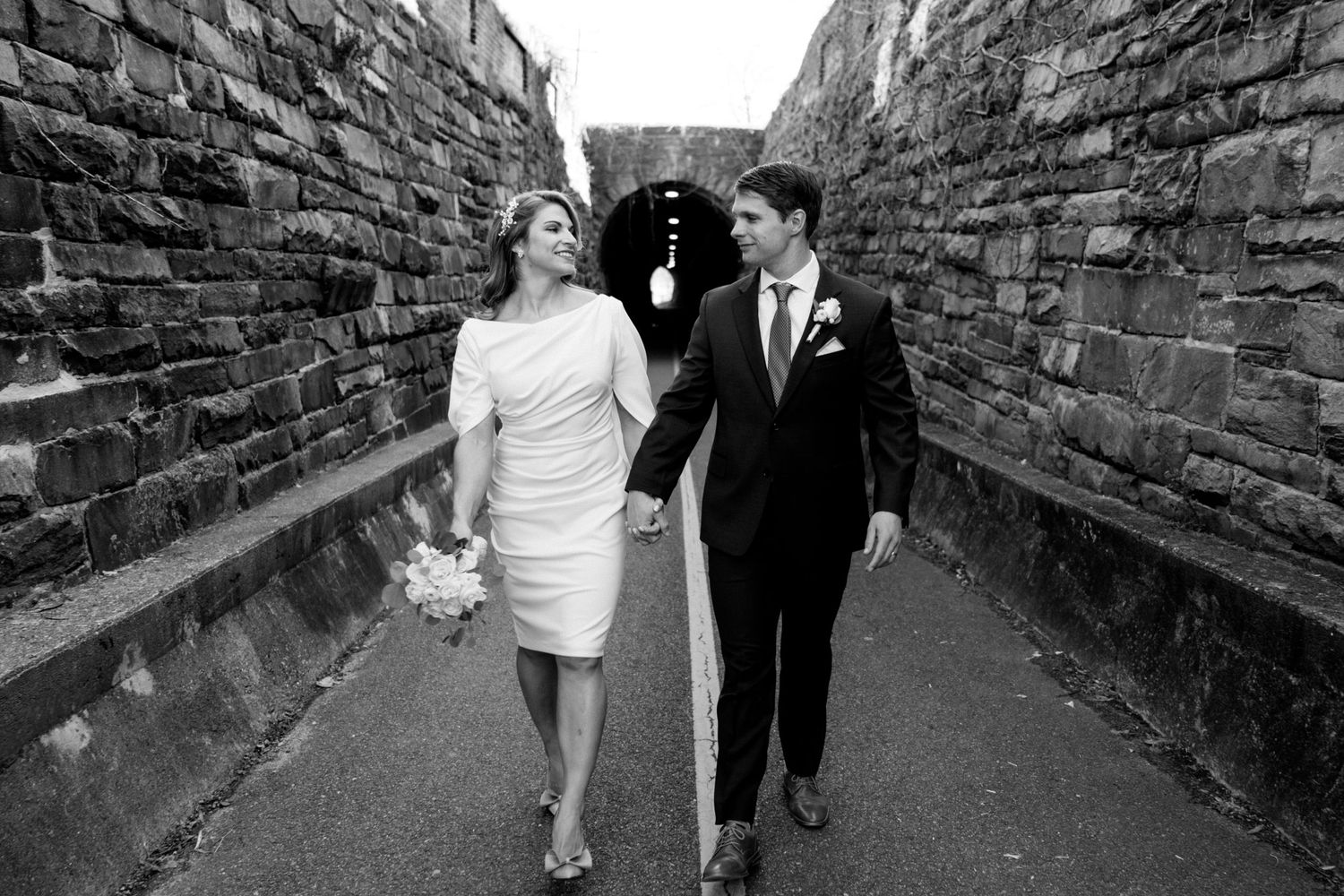 A happy couple walks hand in hand between stone walls in black and white wedding photography.