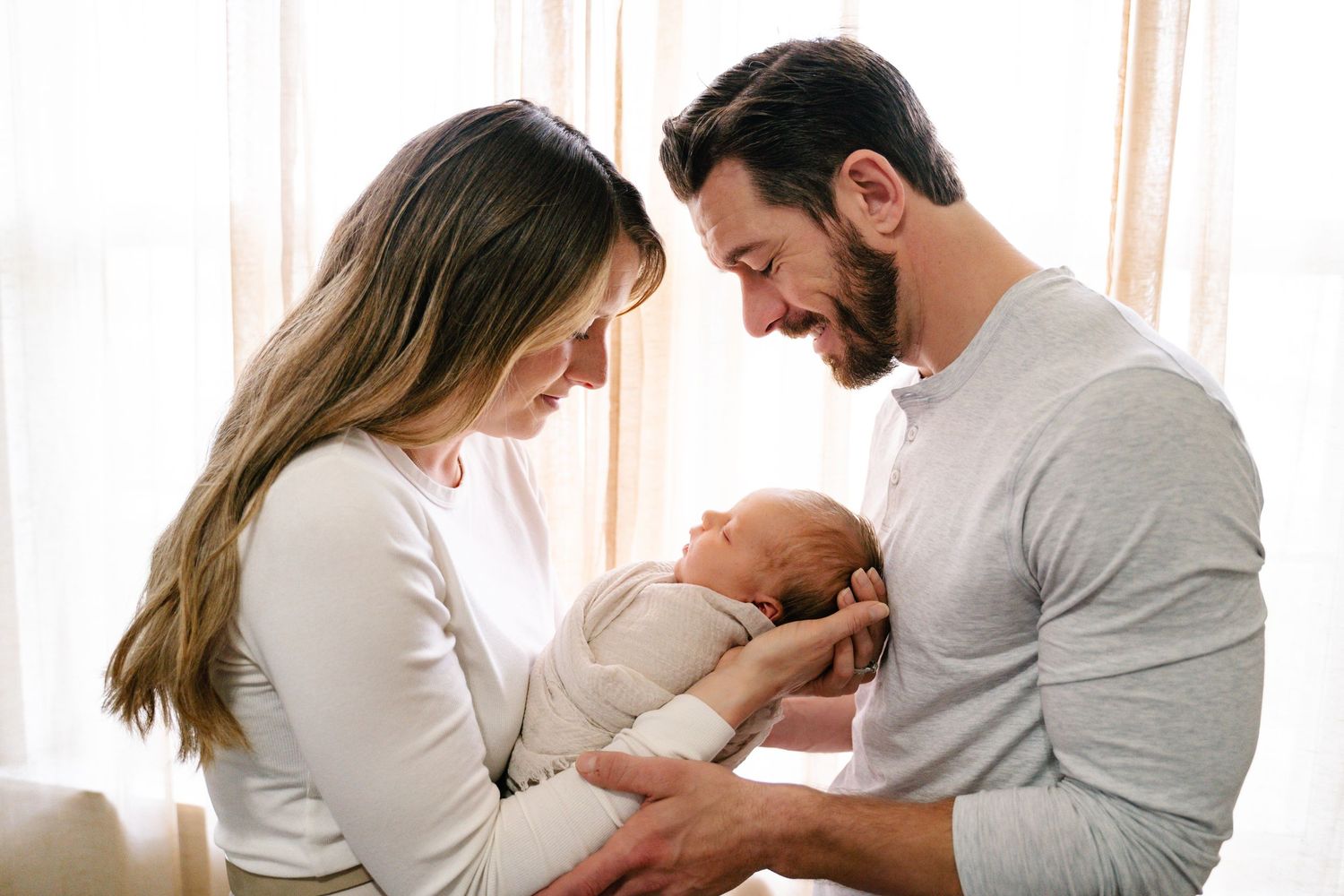 Parents lovingly cradle their newborn baby while sharing an intimate family moment by a window.