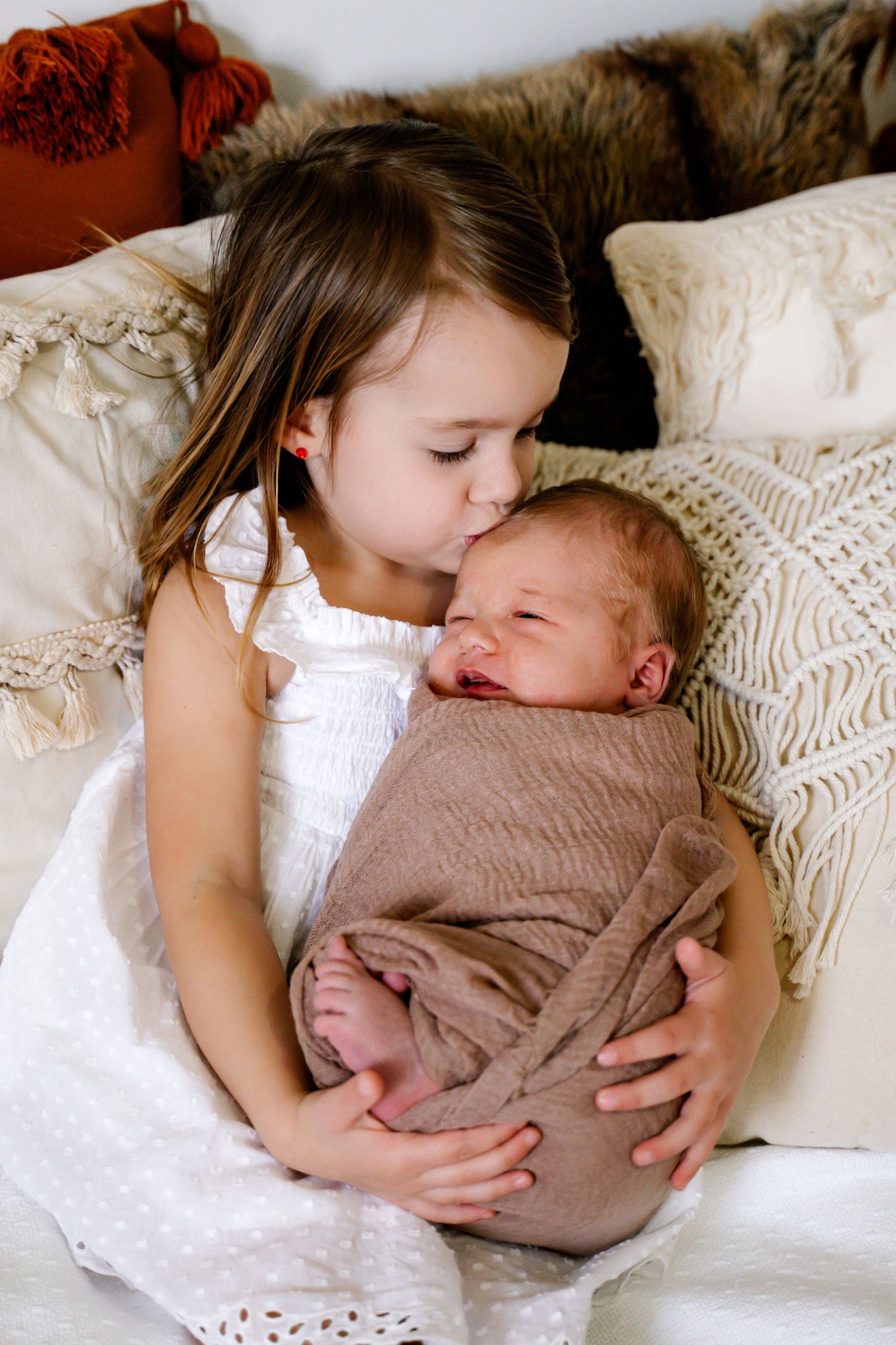 Young girl in white dress cuddles with newborn baby on textured bedding with pillows.