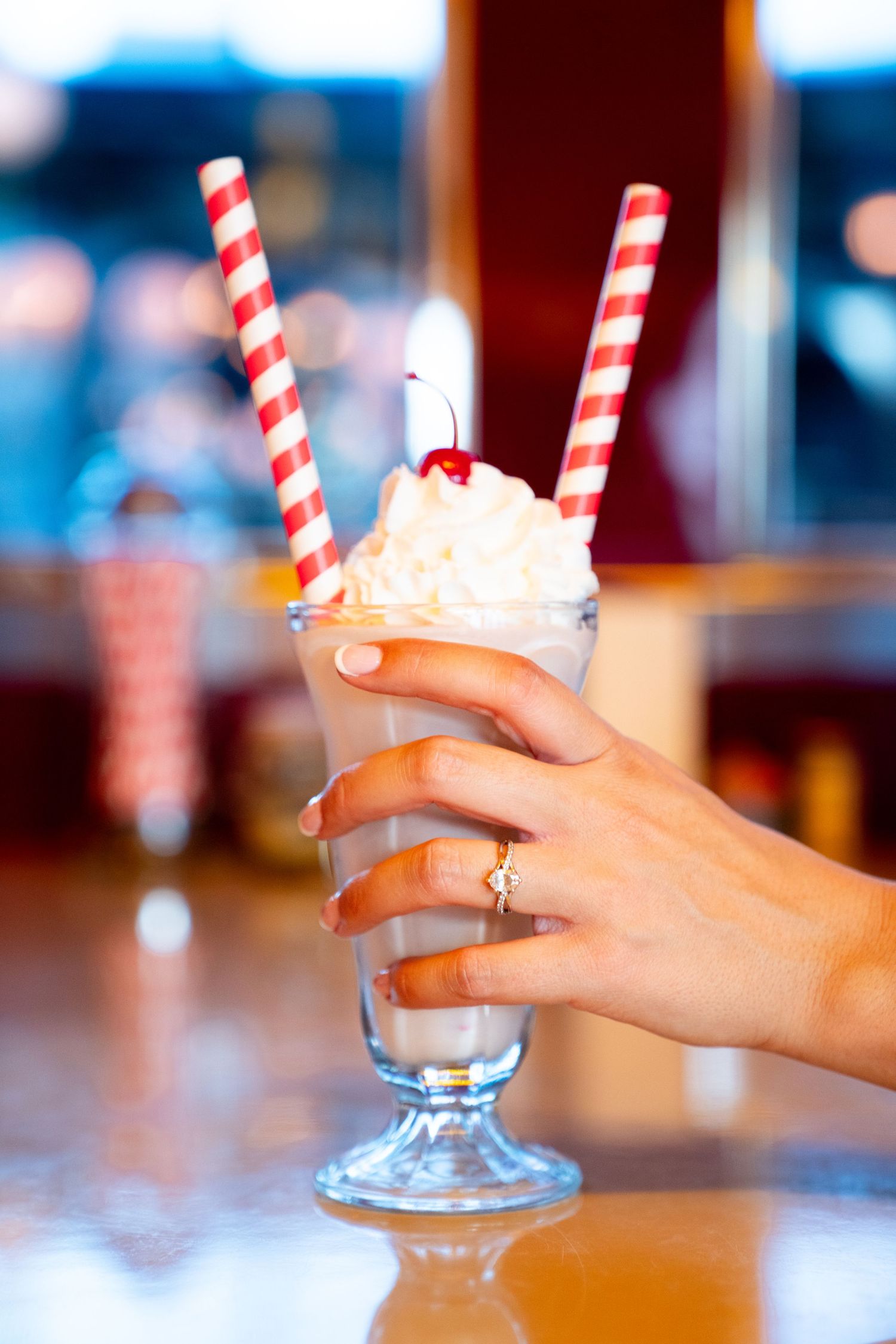 Milkshake with whipped cream and red striped straws served in a classic glass, with a hand wearing an engagement ring. Jessica Paige Photo.