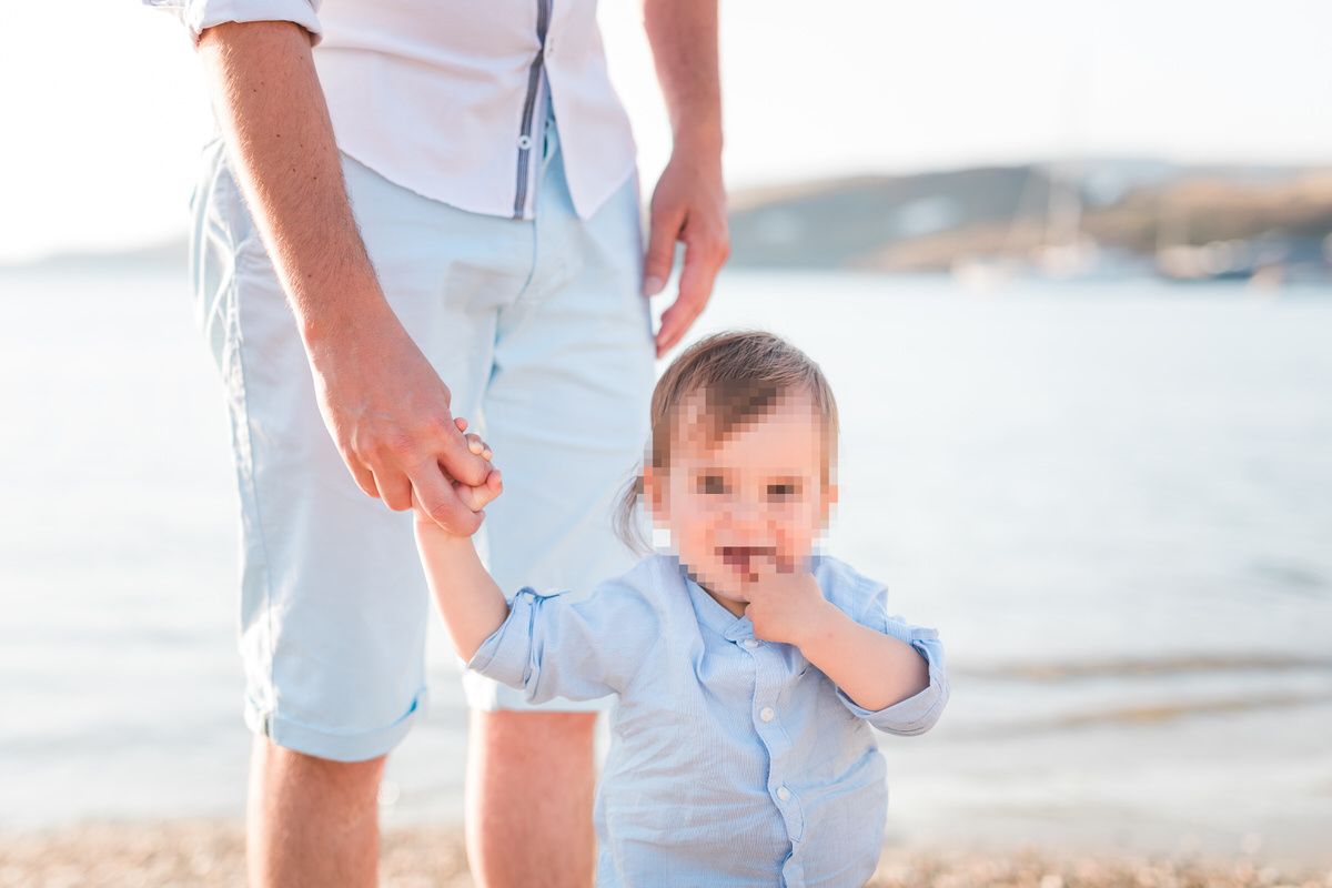 A parent holds a small child's hand while walking along a sunny beach with water in the background.