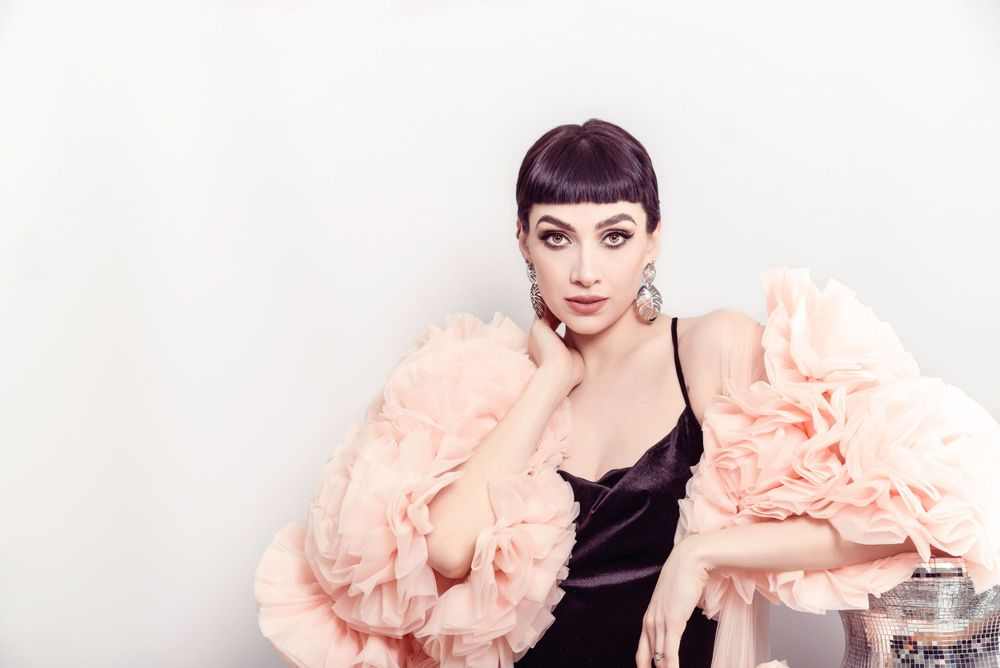 Glamorous figure in black slip dress and peach fur coat poses dramatically against white backdrop.