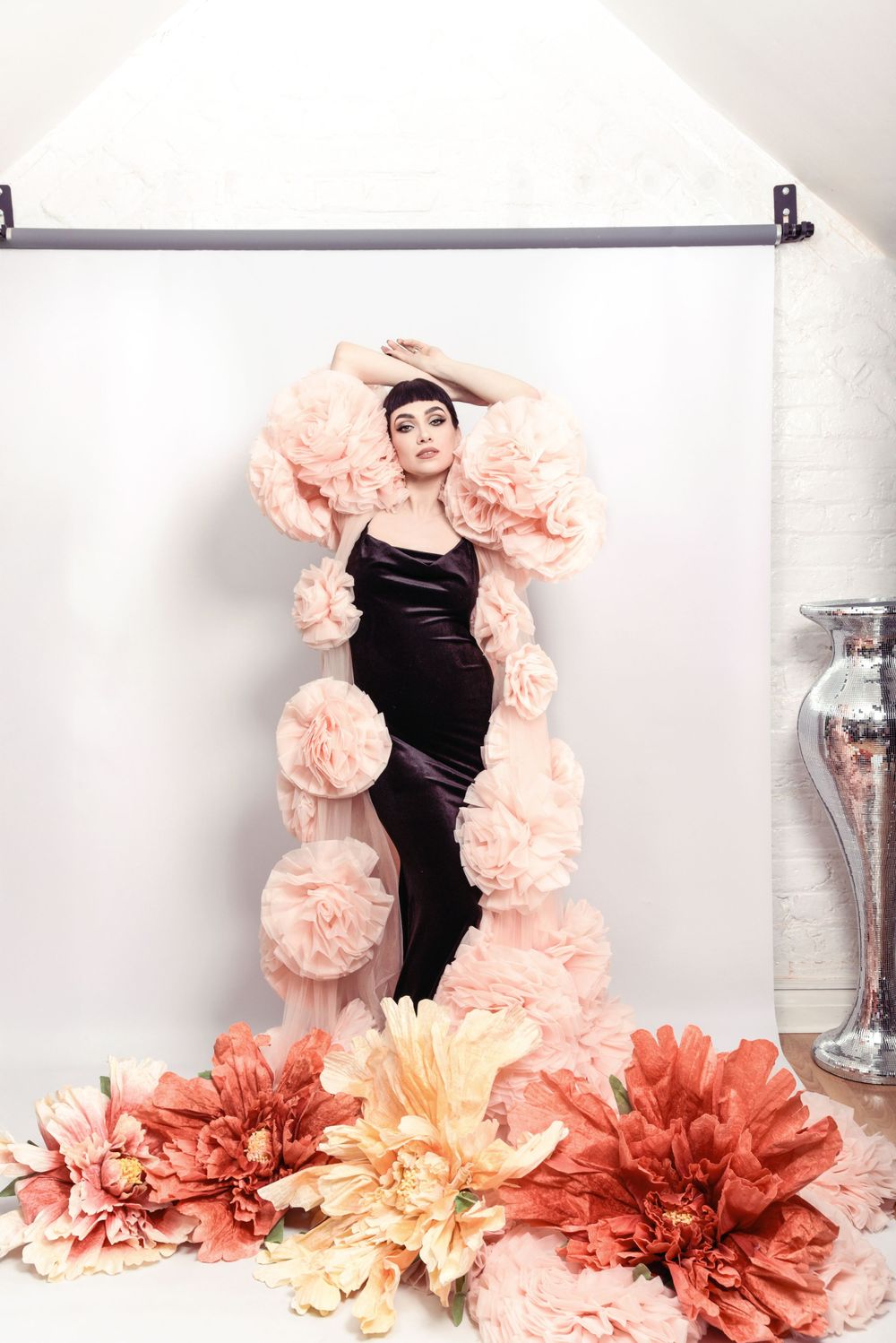 A portrait against white backdrop features a black evening gown adorned with dramatic peach ruffles and coral floral decorations.