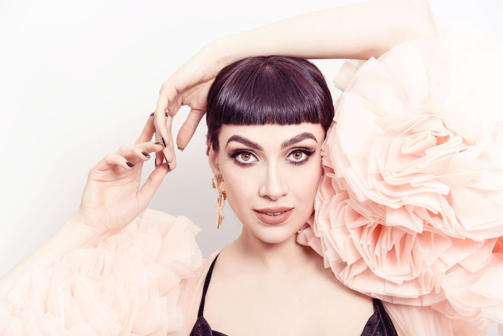 Portrait with styled bangs and dramatic eye makeup against flowing peach-colored fabric in soft lighting.