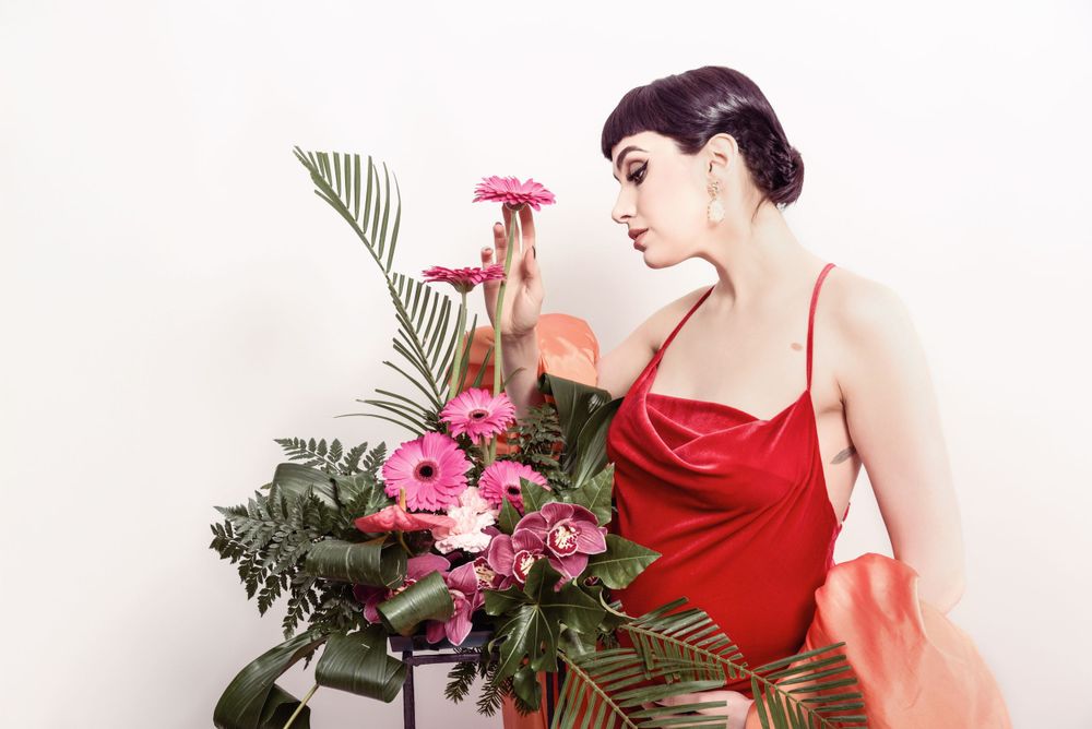 A figure in a red silk slip dress poses with pink flowers and tropical palm leaves against a white background.