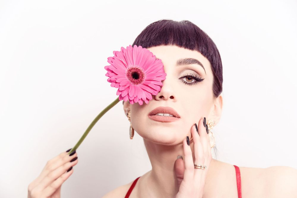 Someone holding a pink gerbera daisy flower against their face with dramatic makeup and short bangs.