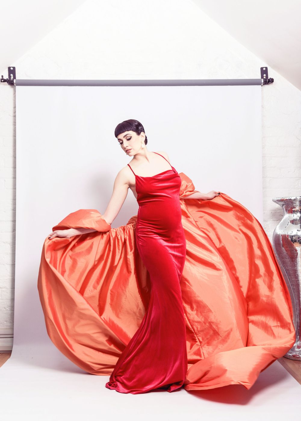 Fashion model in flowing red satin gown poses dramatically against white backdrop with billowing fabric detail.