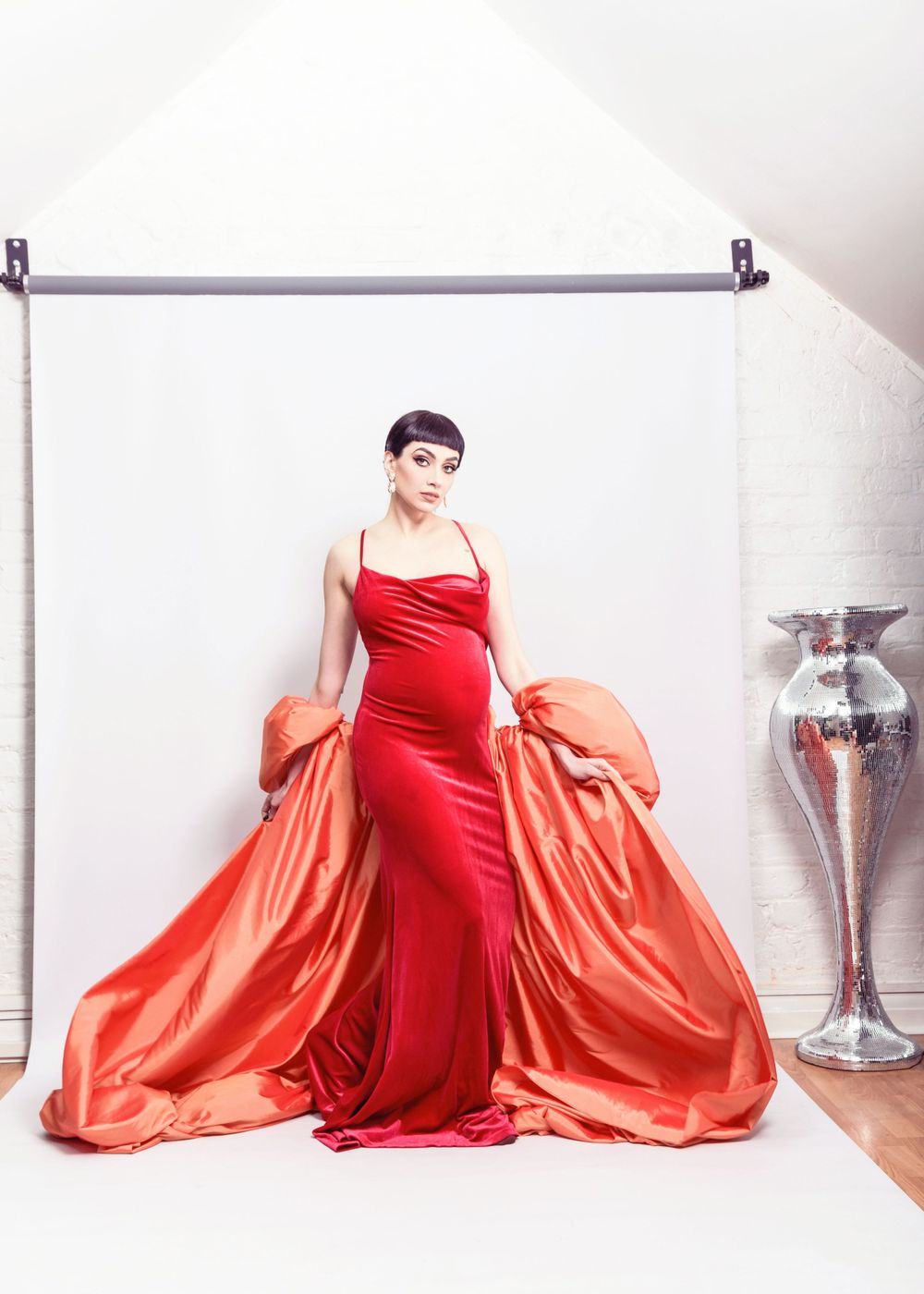 A model poses in a dramatic red evening gown with a flowing coral-colored cape against a white backdrop.