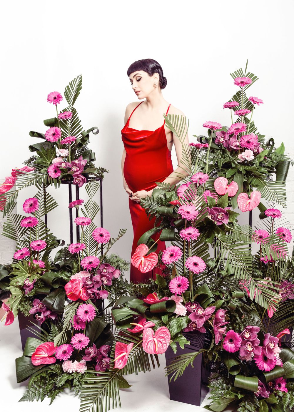 Elegant figure in red dress poses amid vibrant pink tropical flowers and green foliage in studio setting.