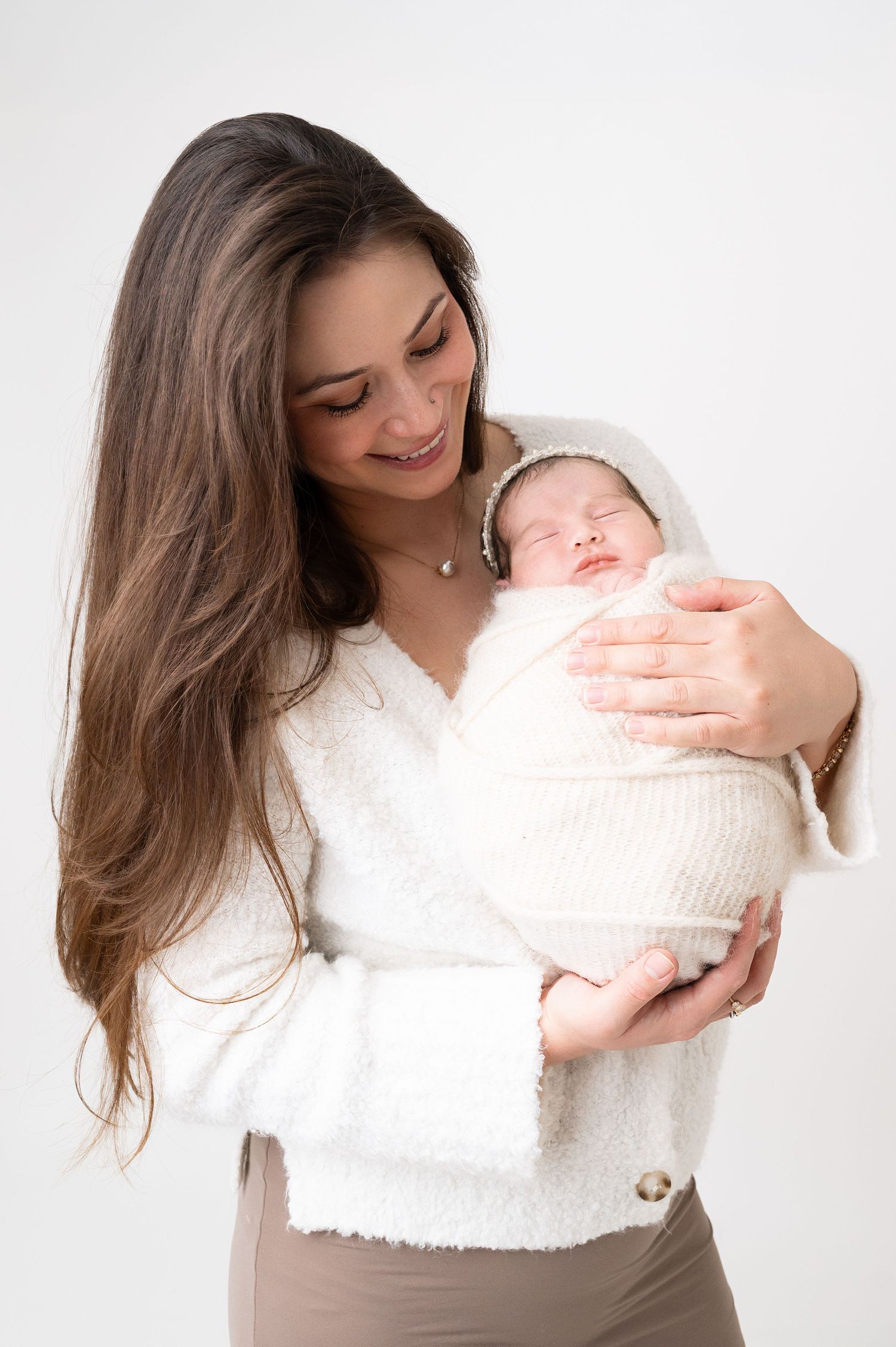 A woman in a white sweater holds a swaddled newborn against a plain white backdrop.