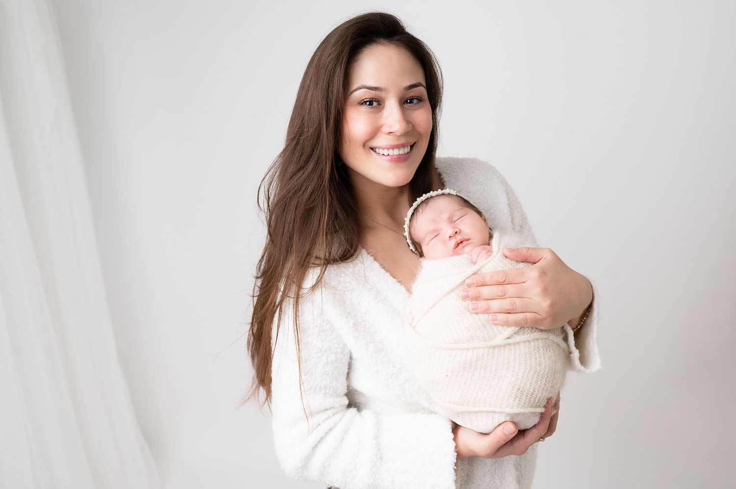 A woman in white holds a sleeping newborn swaddled in cream fabric against a white background.
