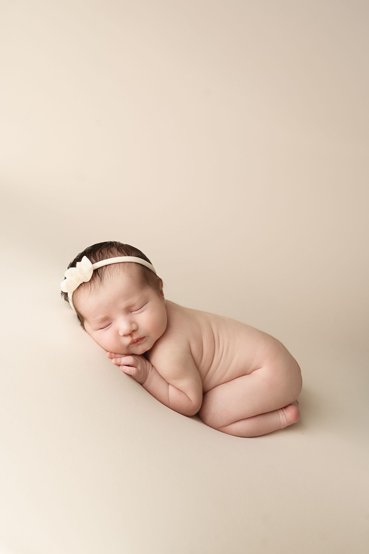 Peaceful sleeping newborn posed on minimalist cream backdrop wearing delicate headband.
