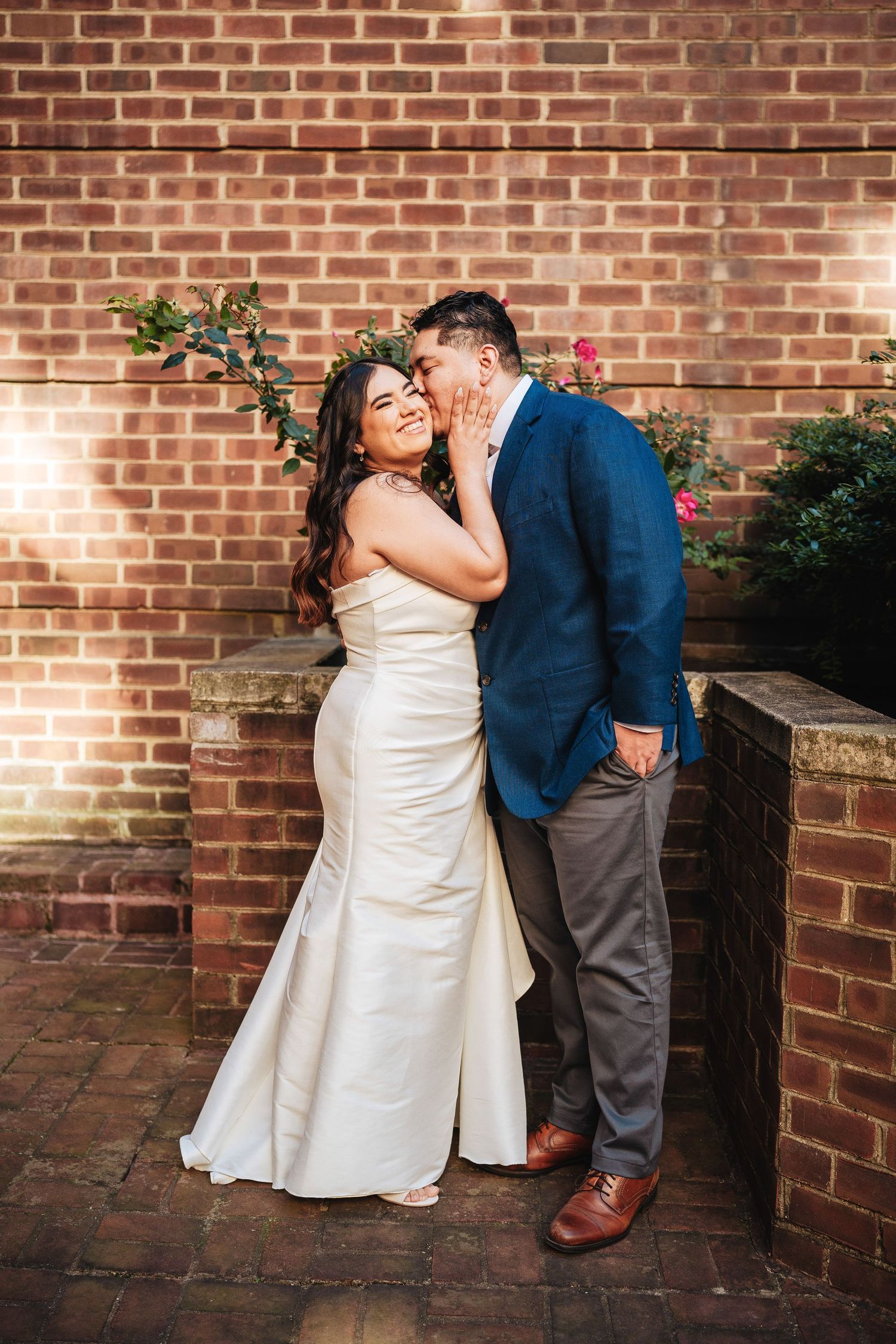 A romantic wedding moment captured against a rustic brick wall with twinkling lights and greenery in the background.