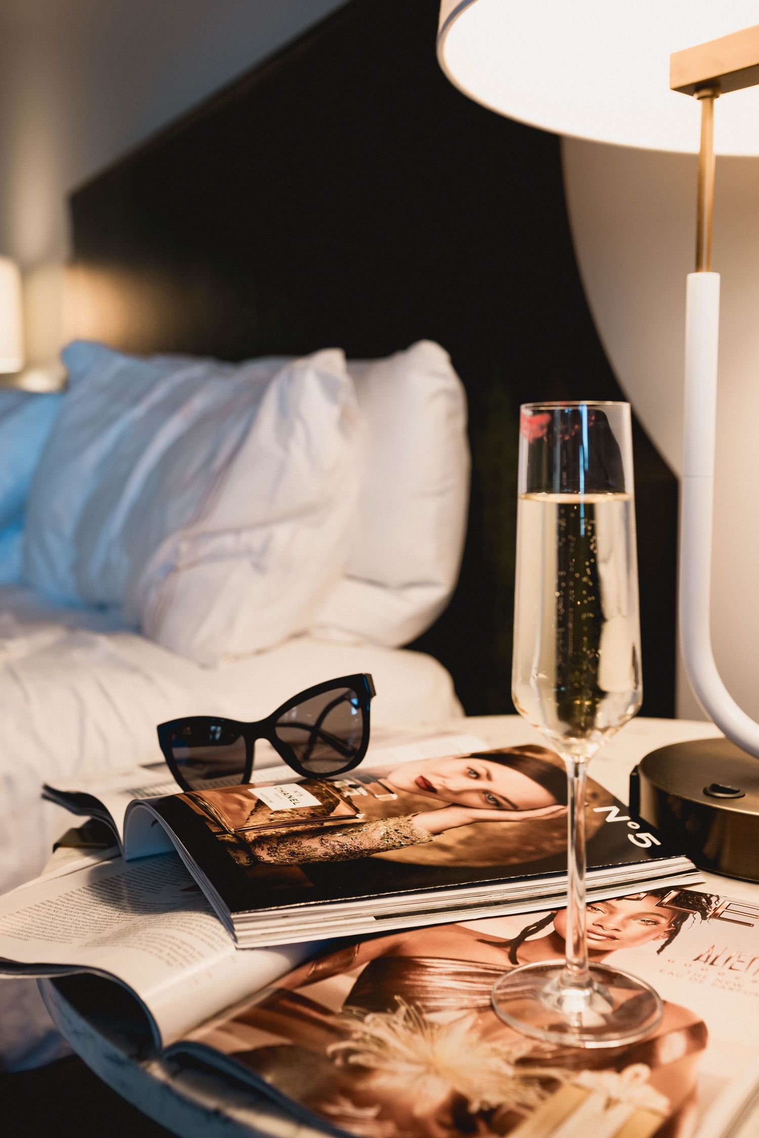 Still life with champagne glass, sunglasses and magazines on white hotel bedding.