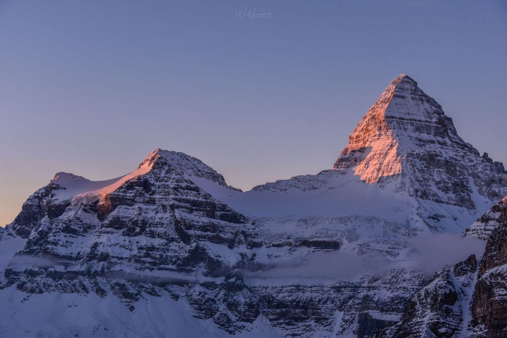 Mountain peaks bathed in alpenglow during sunset with layered rock formations covered in snow.