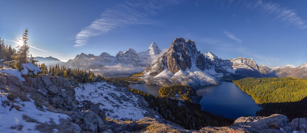 Panoramic mountain landscape with snow-dusted peaks reflecting in alpine lake surrounded by evergreens.
