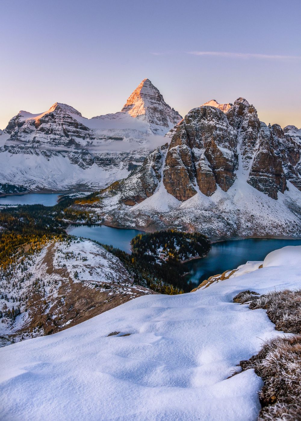 Snow-capped mountain peaks rise majestically over a turquoise alpine lake in the Canadian Rockies during sunset.