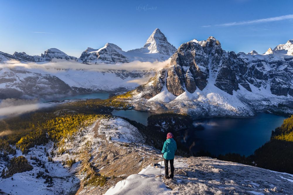 A hiker stands on a snowy viewpoint overlooking Mount Assiniboine and alpine lakes in the Canadian Rockies.