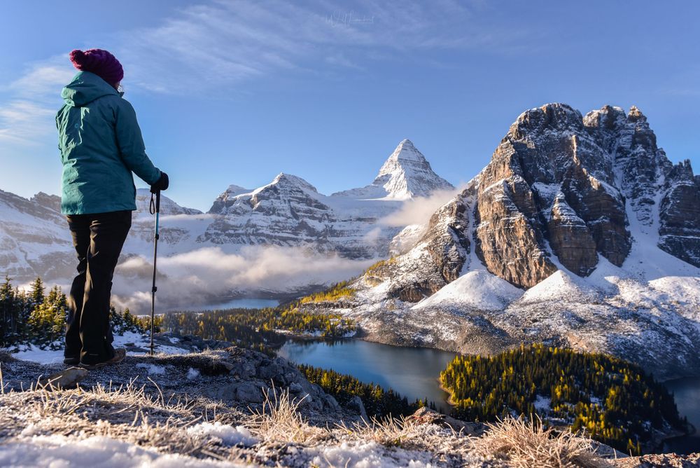 A lone hiker in teal jacket overlooks Mount Assiniboine and pristine alpine lakes on a snowy morning.