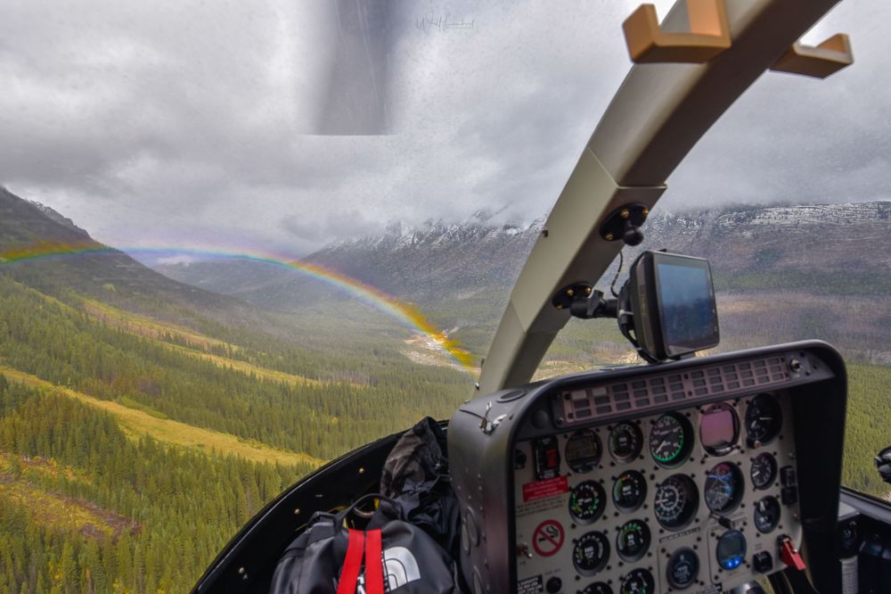 Helicopter cockpit view capturing a rainbow over autumn-colored valleys in the Canadian Rockies.