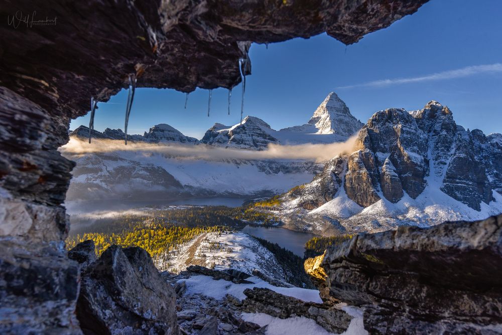 Ice-framed view of Mount Assiniboine and valley through a natural cave opening in winter.