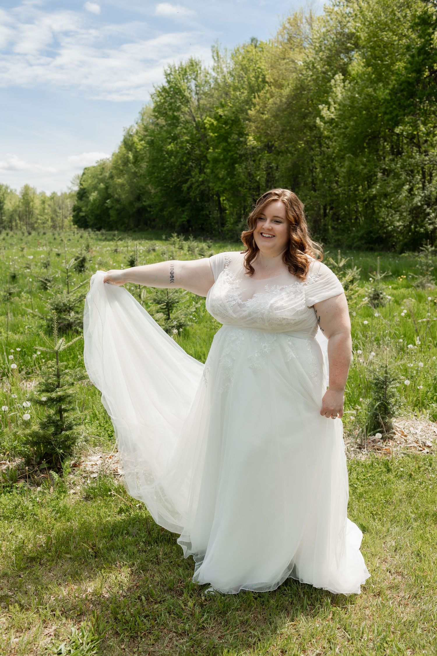 Beautiful wedding dress with flowing tulle skirt and lace cap sleeves photographed in a sunny meadow with green trees.