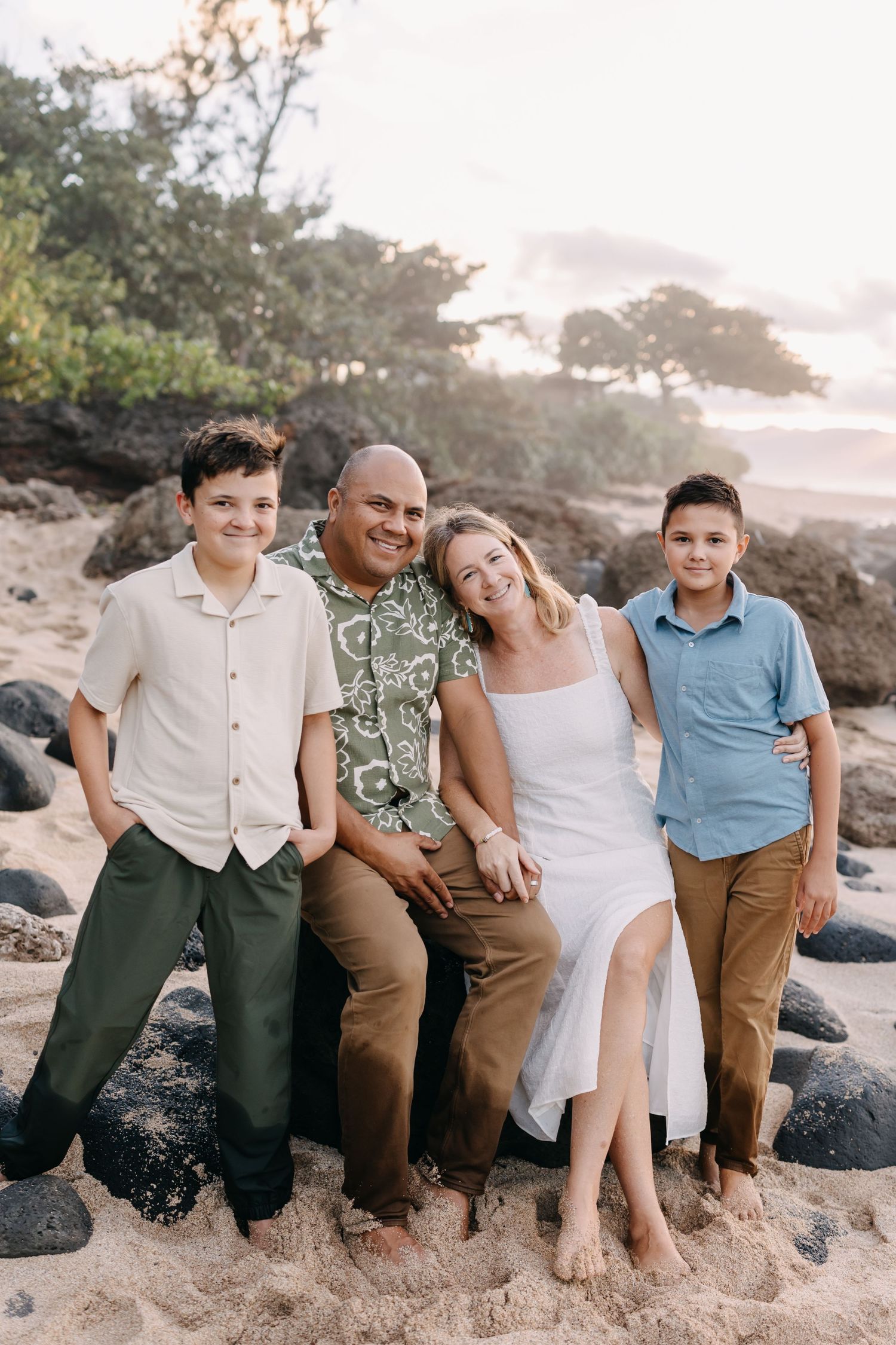 Family of four sitting together on a rocky beach at sunset wearing casual beachwear and Hawaiian shirt.