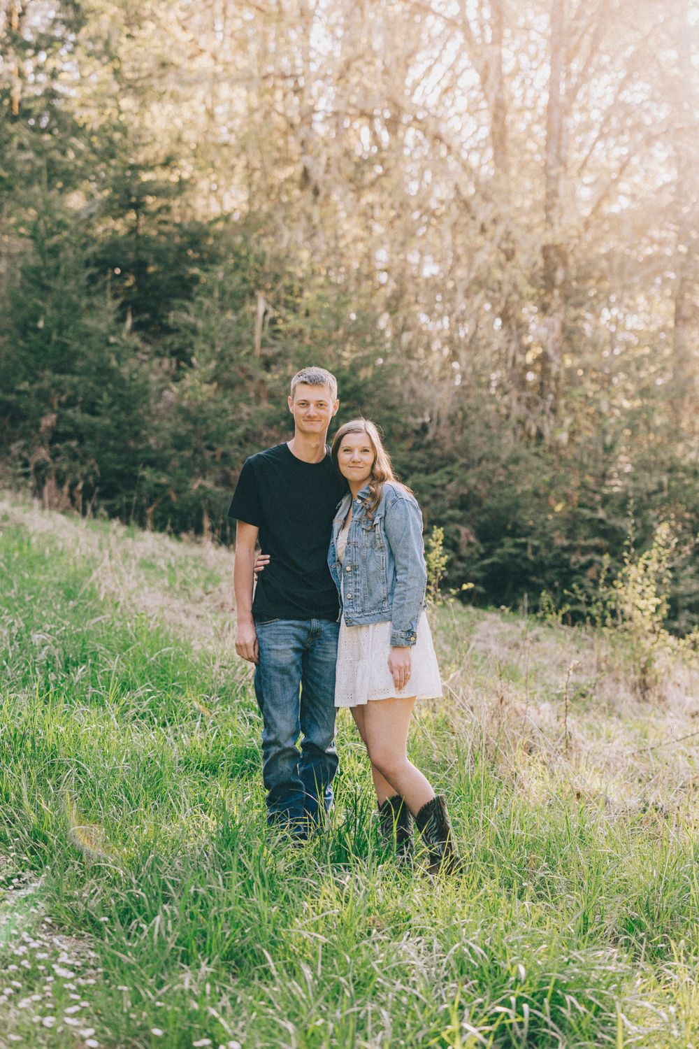 A pair stands together on a grassy hillside with trees in the background during a sunny afternoon.