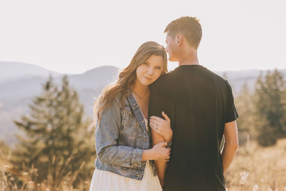 A couple embraces while standing in a mountain meadow on a sunny afternoon with scenic views in the background.