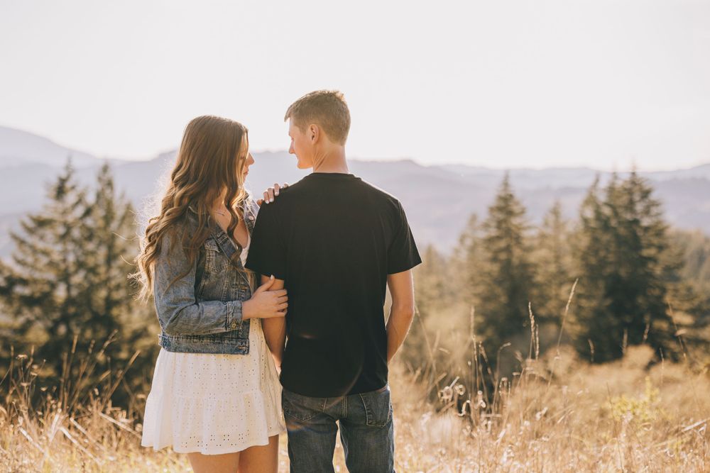 A couple standing in a golden field with mountains and evergreen trees in the distance at sunset.